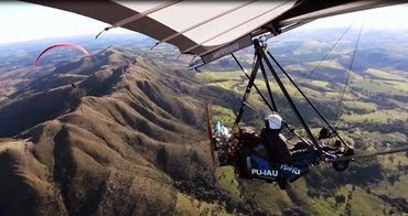 A person flying a microlight aircraft over mountainous terrain with a paraglider in the distance.