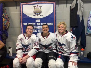 Three female hockey players in white jerseys sitting and smiling in a locker room.