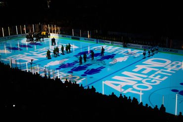 Hockey teams lined up on ice with "AMEND TOGETHER" projection during ceremony.