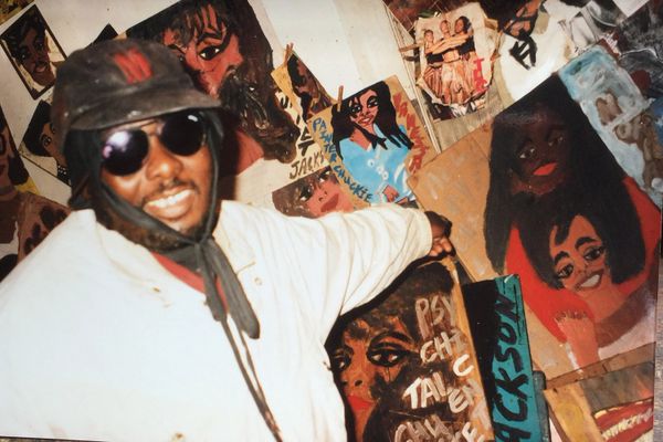 Man in sunglasses and hat posing with colorful paintings of faces.