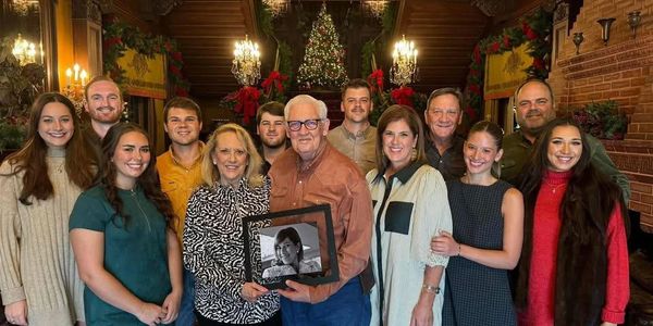 A large family gathers indoors with holiday decorations, holding a framed photo of a woman.