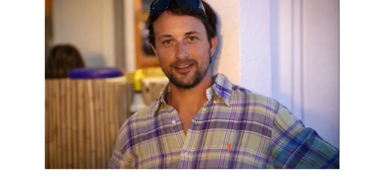 Man in plaid shirt leaning against wall, smiling.