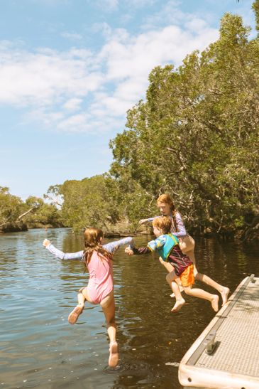 Three kids joyfully jump off a dock into a calm river surrounded by trees.
