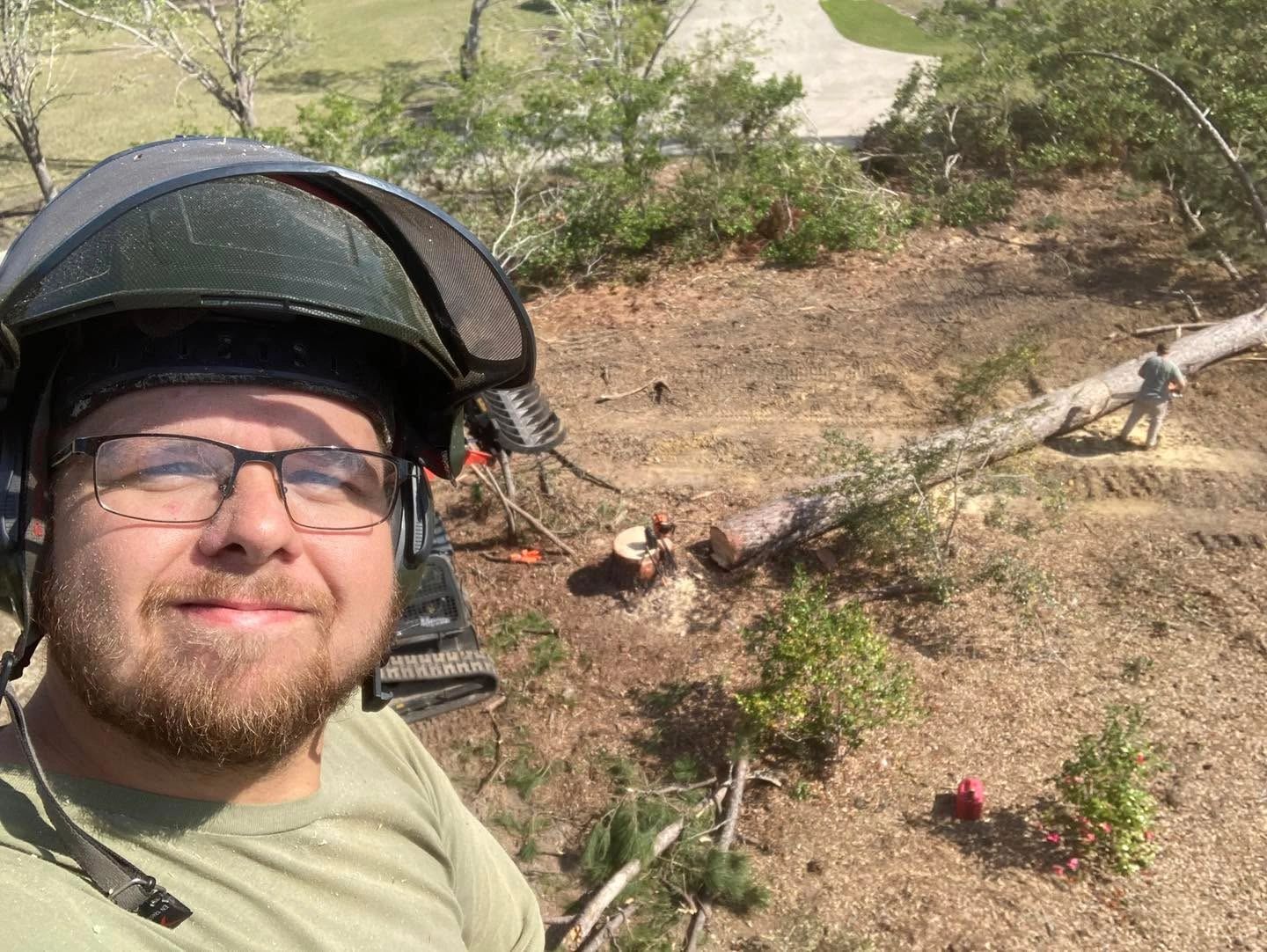 Man wearing safety gear takes selfie while tree cutting work is underway below.