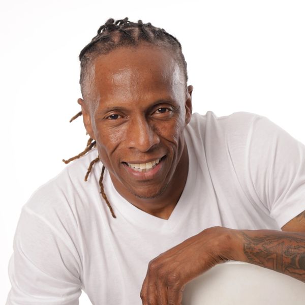 Smiling man with braided hair and tattoo, wearing a white shirt.