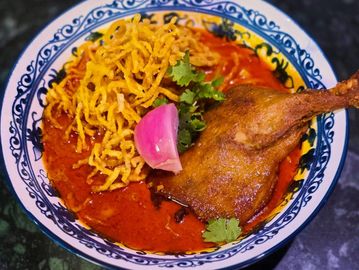 A bowl of spicy duck curry with crispy noodles and garnishes.