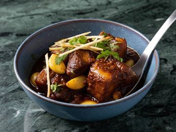 Spicy cooked meat garnished with garlic cloves, ginger, and cilantro in a blue bowl.