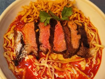 Bowl of spicy red curry noodles topped with sliced medium-rare beef and crispy fried noodles.