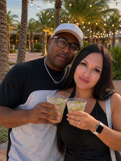 Couple enjoying drinks outdoors with palm trees and lights in the background.