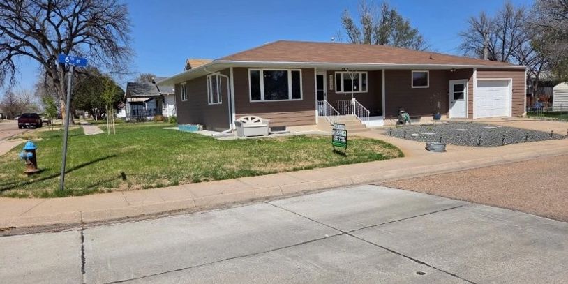 Single-story brown house on a corner lot with a green lawn and a for sale sign.