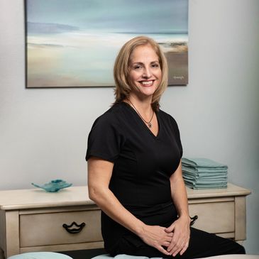Smiling woman in black scrubs sitting on a massage table in a calming room.