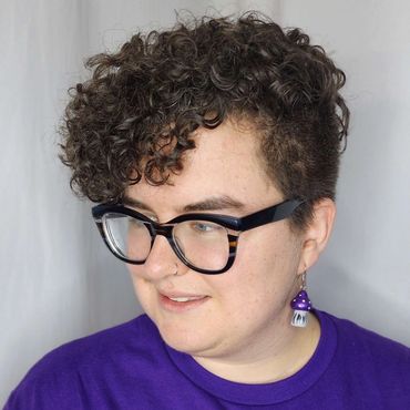Person with curly hair, glasses, and a purple shirt featuring a colorful graphic and mushroom earrings.