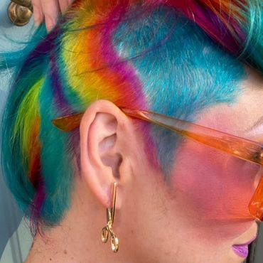 Person with vibrant rainbow-colored hair and gold scissor-shaped earrings.