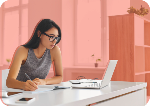 A woman in glasses writes notes in a notebook beside her laptop, representing shaping the vision.