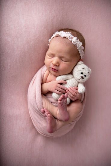 newborn girl wrapped in pink with pink headband and clutching a white knitted teddy bear