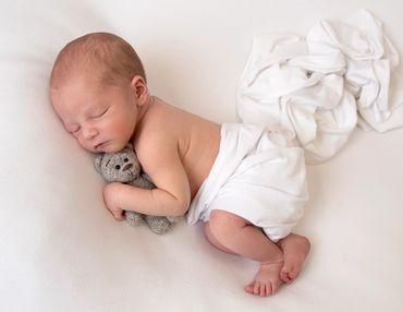 newborn asleep on his side cuddling a grey knitted teddy bear on white