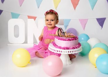 Smiling baby girl in pink dress celebrates first birthday with cake and balloons.
