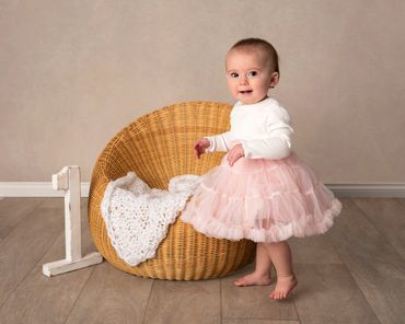 Adorable baby girl in a pink tutu dress standing next to a wicker chair.