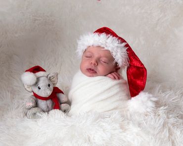 baby boy asleep swaddled in white wearing a santa christmas hat with a small christmas elephant