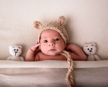 wide eyed newborn tucked in a bed wearing a knitted bear hat, with 2 knitted teddy bears