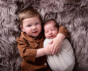 toddler and his newborn swaddled sister cuddling on grey fur