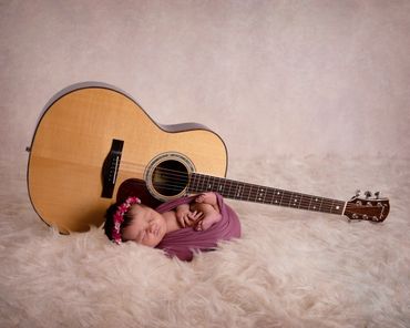 newborn girl swaddled in a burgundy wrap and wearing a flower headband with a guitar on a cream fur