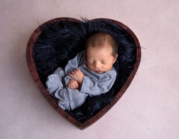 newborn boy dressed in a blue onesie on navy fur in a dark wood heart bowl