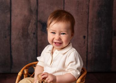 Smiling baby in a cream sweater sitting on a wicker chair against a wooden background.