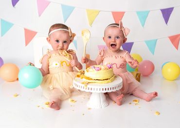 Two babies in pastel dresses enjoy smashing a cake with colorful balloons and bunting in the background.