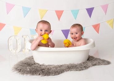 Two babies playing with yellow rubber ducks in a white tub with colorful bunting in the background.