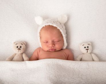 sleeping newborn in a white bear hat with two knitted teddy bears