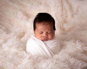 sleeping newborn baby with lots of hair swaddled in potatoe sack pose on cream fur