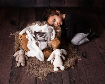 beautiful toddler crouching next to some wood with a baby scan photo and some cuddly teddies