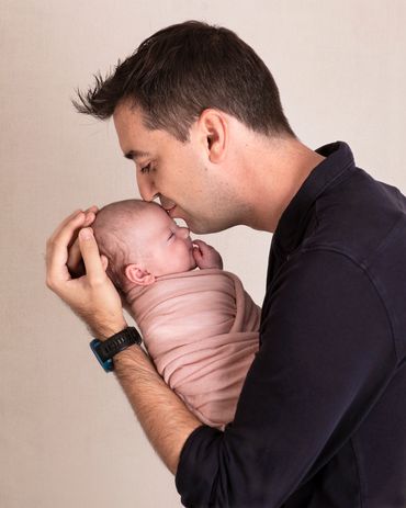 father kissing his newborn daughter's head while she smiles and is swaddled in a peach wrap