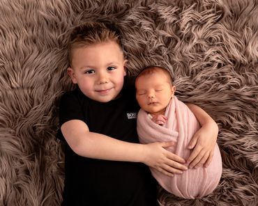 4 year old holding his newborn sister who is swaddled in peach and asleep on brown fur
