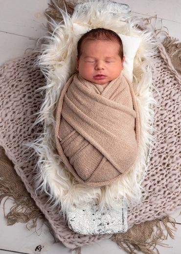sleeping newborn swaddled in a knitted wrap on a bed of white fur in a trench bowl on a blanket