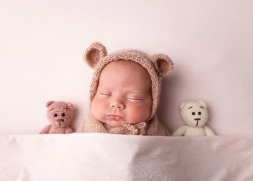 sleeping newborn tucked in bed wearing a knitted pink teddy bear hat with two knitted teddy bears