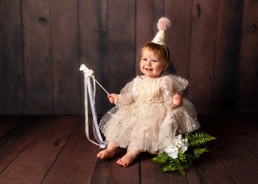 Smiling baby in a delicate dress and party hat holding a ribbon wand.