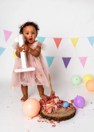 A toddler in a pink dress holds a large number one with a smashed cake and balloons around.