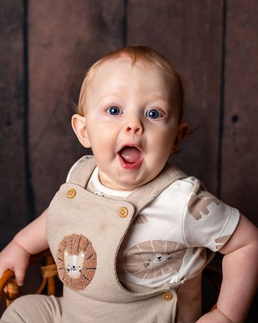 Surprised baby with blue eyes wearing beige overalls with a lion patch.