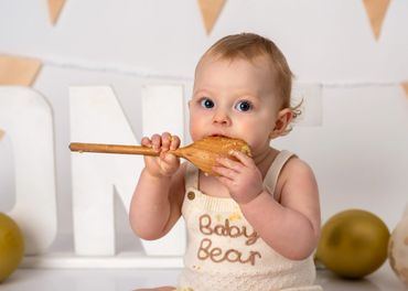 Baby in a cream outfit tasting cake off a wooden spoon.