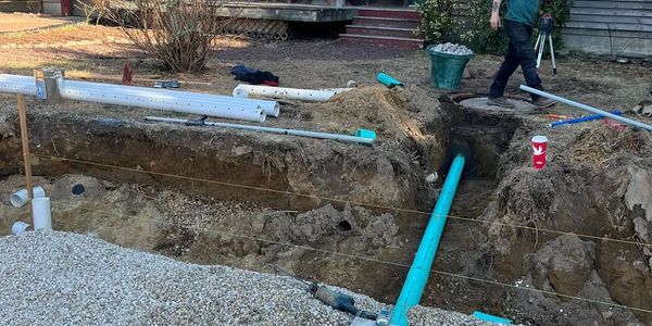 Man working on outdoor plumbing near a house under clear sky.