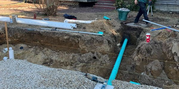 Man working on outdoor plumbing near a house under clear sky.