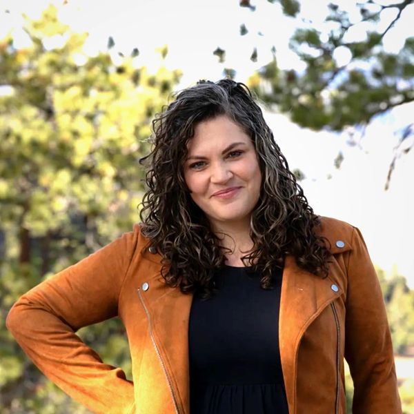 A woman with curly hair wearing a brown jacket and smiling outdoors.