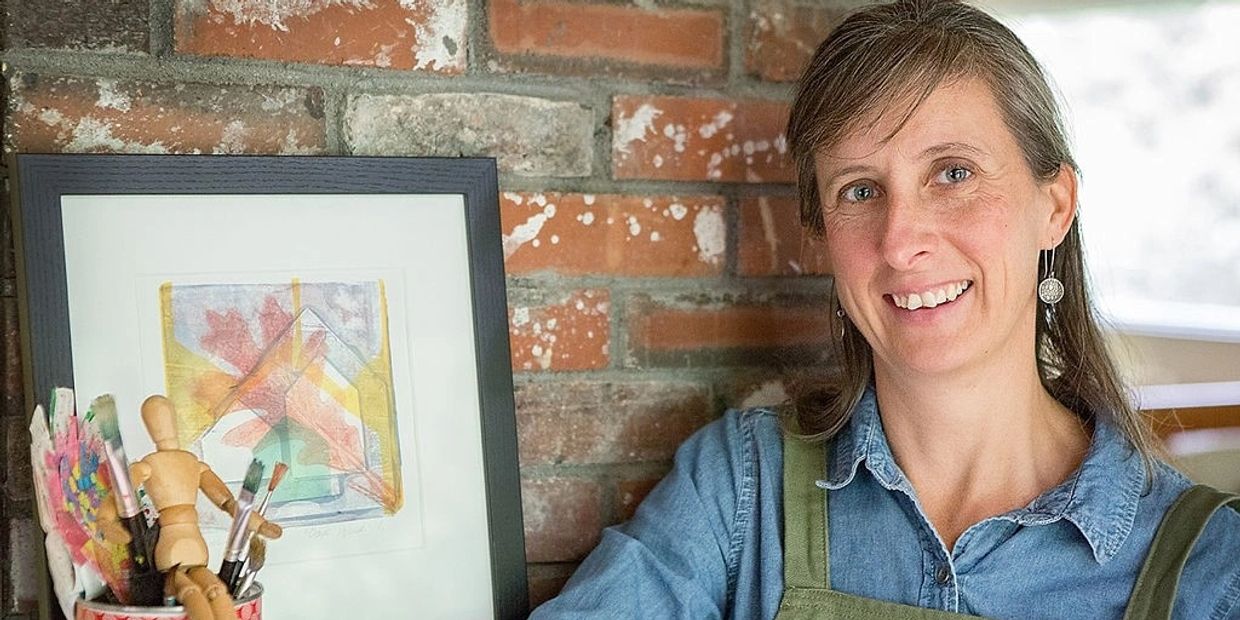 Woman artist smiling beside her framed artwork and brushes.