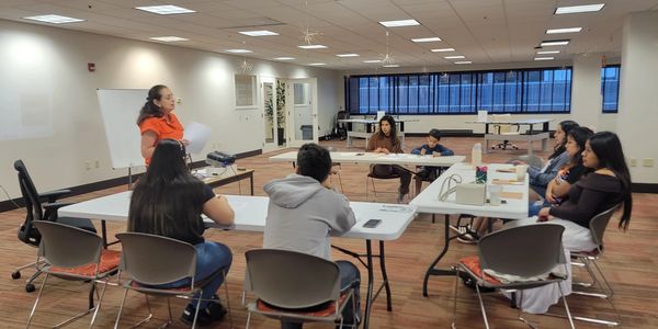 A woman in orange leads a meeting with people seated around tables in a large room.