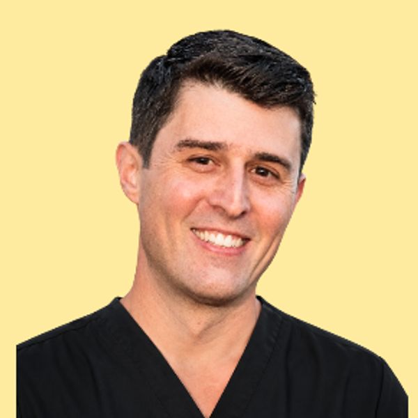 Smiling man in black medical scrubs against a yellow background.