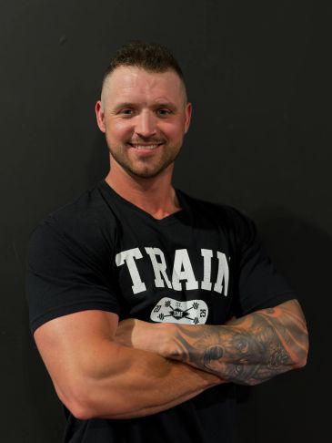Smiling muscular man with tattooed arm wearing a TRAIN shirt.