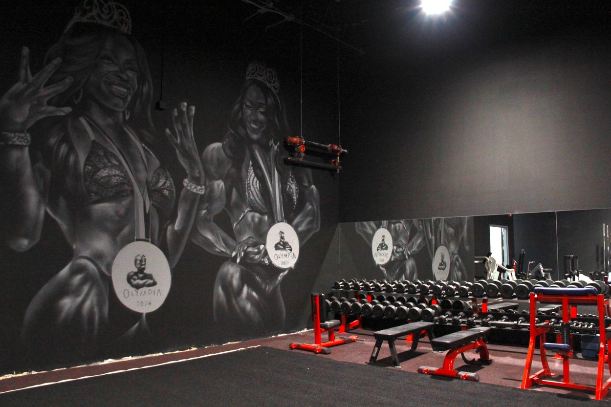 Gym interior with mural of female bodybuilders and red workout equipment.