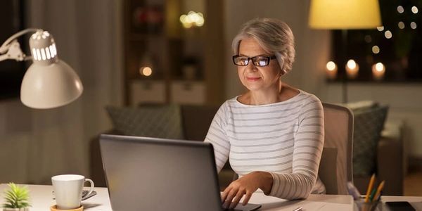 Older woman working on a laptop in a cozy, softly lit room at night.